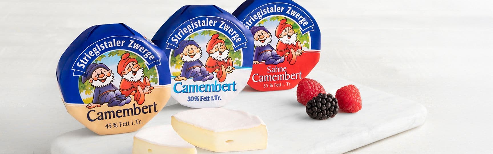 Drei Sorten Striegistaler Zwerge Camembert (45 %, 30 % und Sahne-Camembert 55 %) auf Marmorplatte, dazu Camembert-Stücke und frische Beeren.