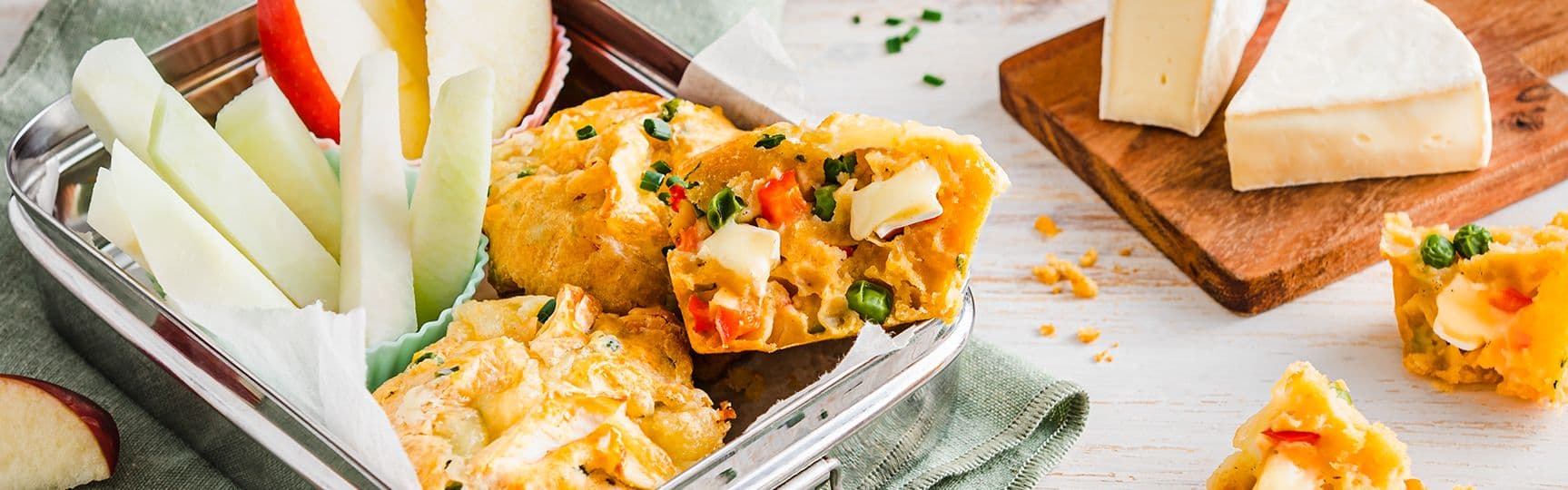 Herzhafte Muffins mit Gemüse und Camembert in einer Brotdose, dazu Apfel- und Gurkensticks, im Hintergrund Camembert-Stücke auf einem Holzbrett.