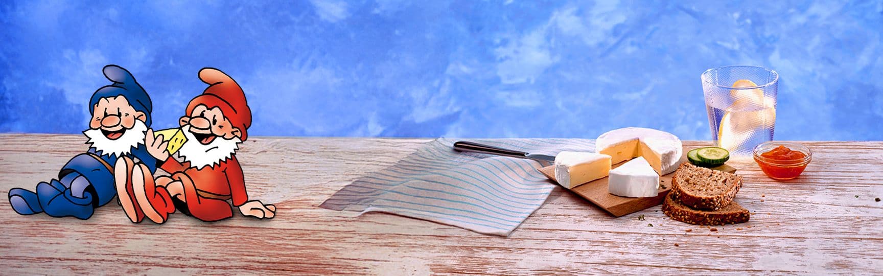 Zwei gezeichnete Striegistaler Zwerge mit Camembert, daneben ein gedeckter Tisch mit Camembert-Stück, Vollkornbrot, Gurkenscheiben, Konfitüre und Getränk mit Zitronenscheiben.